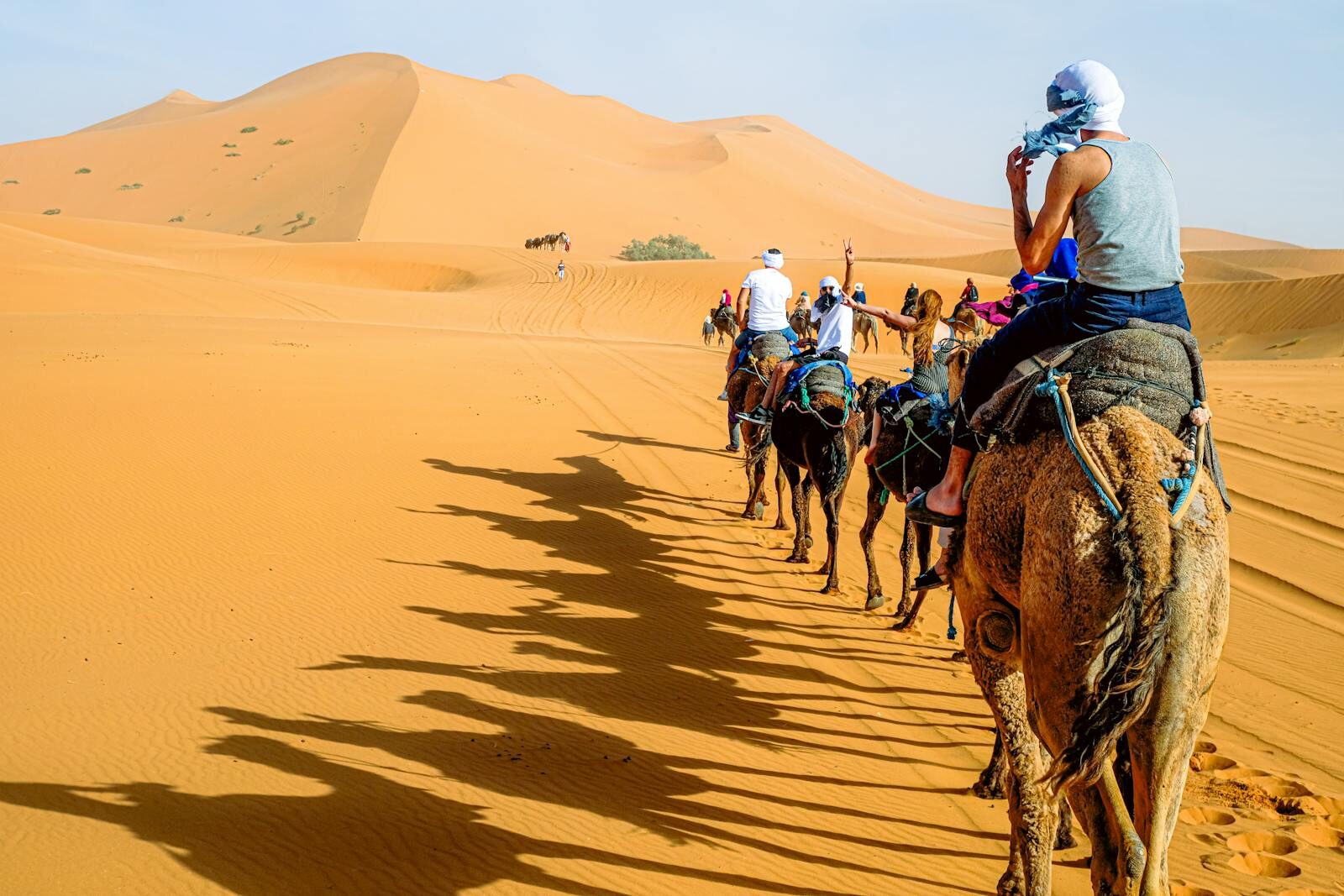 A group of people riding camels across the vast sand dunes under a bright sky.